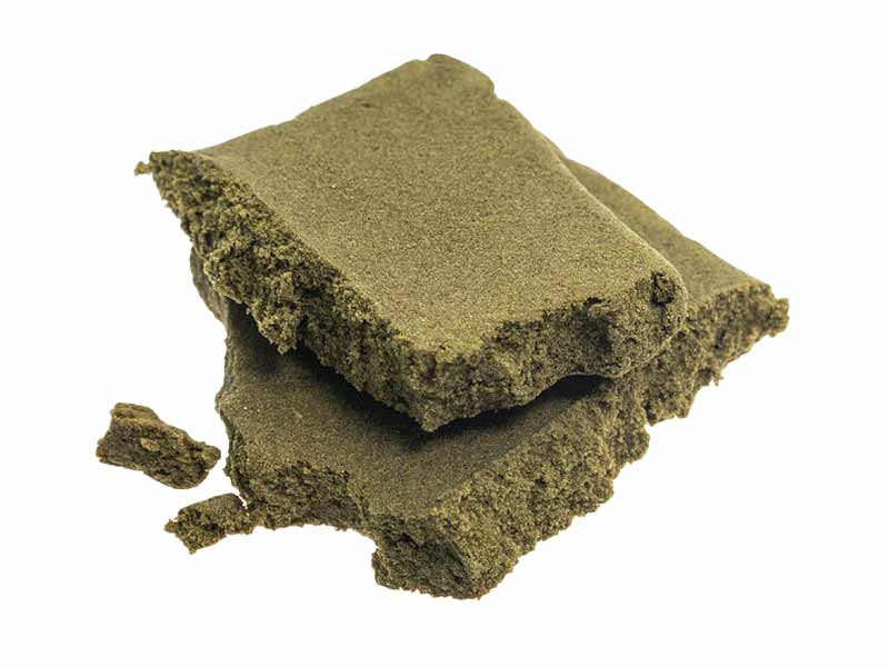 Two rectangular pieces of Moroccan hash with a crumbly, compact texture and brownish‑green color, fragments scattered around.